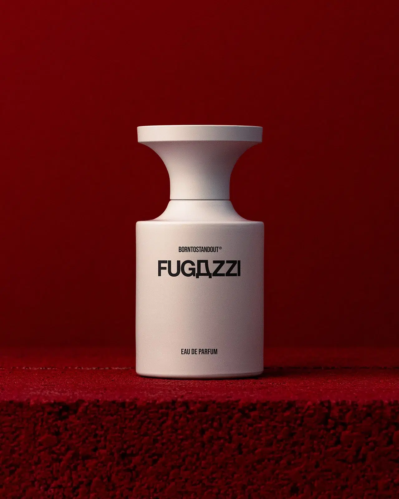 Білий флакон BORNTOSTANDOUT FUGAZZI на бордовому ворсистому подіумі перед темно-червоною стіною