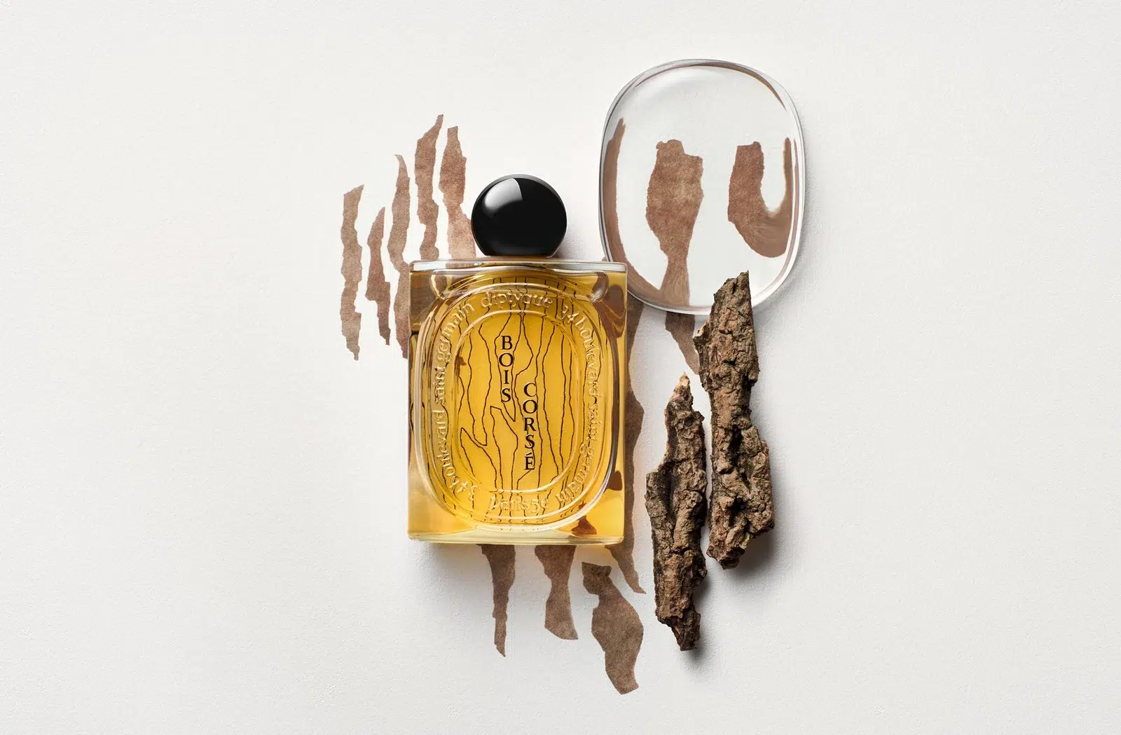 Флакон Diptyque з круглою чорною кришкою поряд із корою та декоративною тацею на світлому тлі